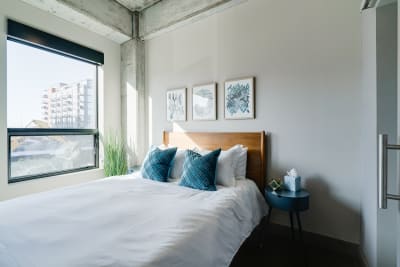 A stylish bedroom with white bedding, turquoise pillows, abstract art, and a window overlooking an urban landscape.
