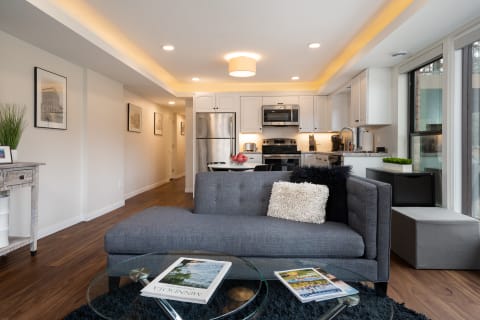 Interior view of a modern living area with a gray sofa, glass coffee table, and a kitchen in the background.