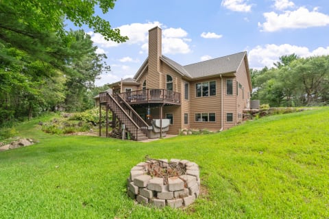 A house with a wooden deck, surrounded by grass and trees, with a fire pit in the foreground.