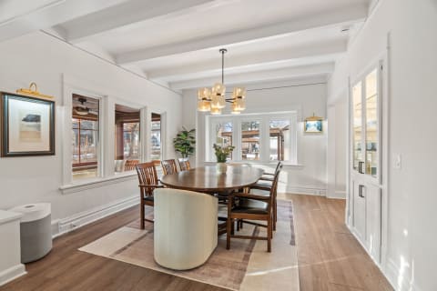 A spacious dining room featuring a wooden table and chairs, modern chandelier, and large windows allowing sunlight in.