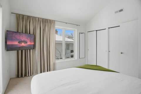 A minimalist bedroom with a wall-mounted TV displaying a sunset, white linens on the bed, and beige curtains.