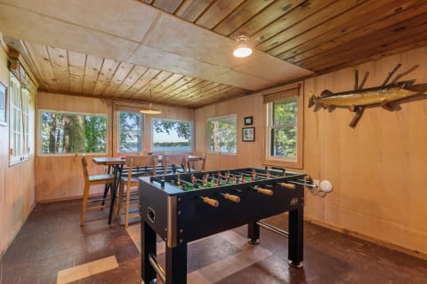 Interior of a game room with a foosball table, wooden decor, and large windows overlooking trees.