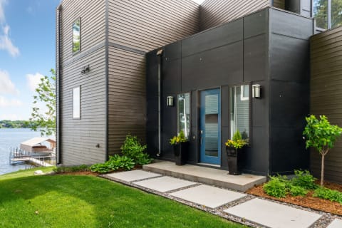 Modern black house entrance by the lake with blue door and lush greenery.