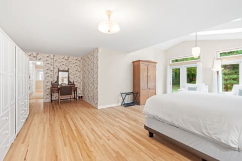 Spacious bedroom featuring hardwood floors, a white bed, an antique wardrobe, and large windows with greenery views.