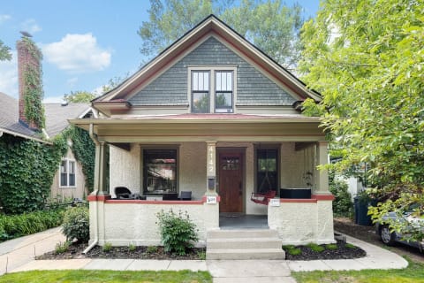 A two-story house with cream stucco, red trim, and a lovely front porch surrounded by greenery.