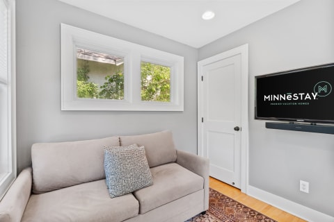 A light gray living room with a beige couch, windows, and a mounted TV displaying the Minnestay logo.