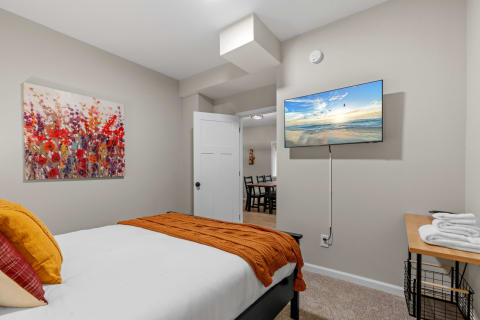 Cozy bedroom featuring a bed with an orange throw blanket and vibrant pillows, a mounted TV displaying a beach scene, and colorful artwork.