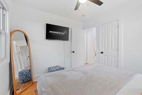 A modern bedroom with a gray bedspread, a wooden-framed mirror, a patterned ottoman, and a flat-screen TV.