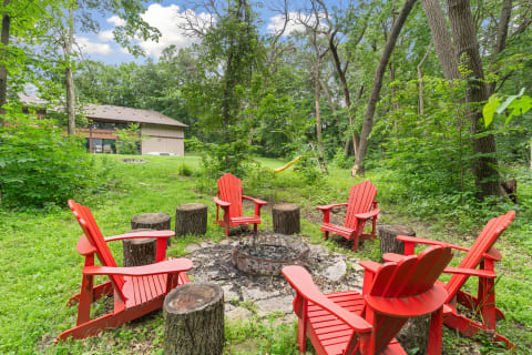 A circular arrangement of red Adirondack chairs surrounding a fire pit in a green wooded area.