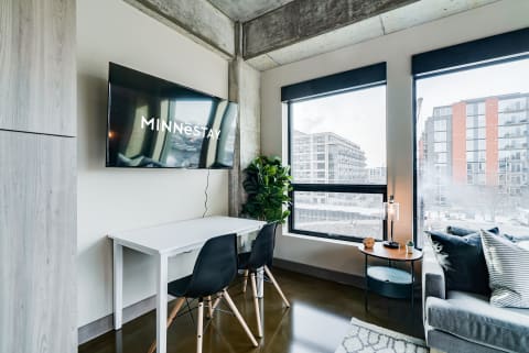 A modern living room featuring a flat-screen TV, workspace, gray sofa, and large windows.