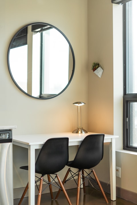 A minimalist workstation with a round mirror, two black chairs, and a desk lamp in a bright room.