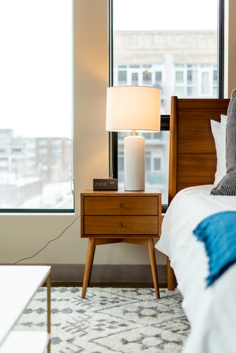 A stylish bedside table with a lamp and clock in a cozy bedroom setting, drizzled windows in the background.