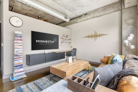 Contemporary living room with a tan leather couch, modern decor, and the MINNēSTAY logo on a flat-screen TV.