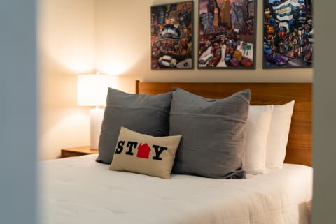 A well-decorated bedroom featuring a bed with plush gray and beige pillows, illuminated by a stylish lamp, and colorful urban art on the wall.