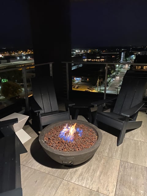 Fire pit on a balcony surrounded by modern chairs with city lights in the background at night.