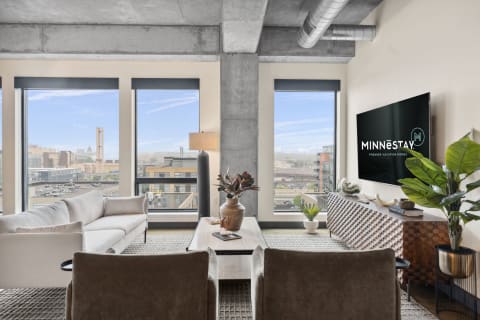 A modern living room featuring large windows, a light sofa, brown armchairs, and city views, with a 'MINNESTAY' logo on the TV.