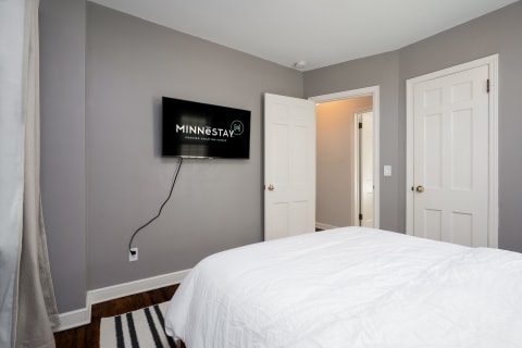 A modern bedroom with a white comforter and a flat-screen TV displaying a logo.