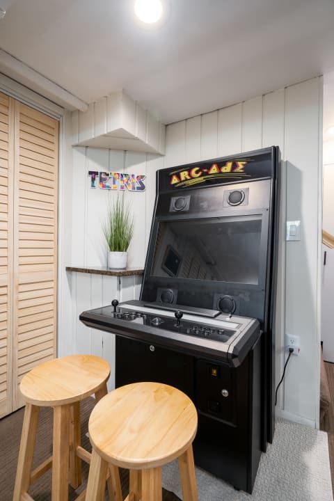 A classic arcade game machine with wooden stools and a plant in a recreational space.
