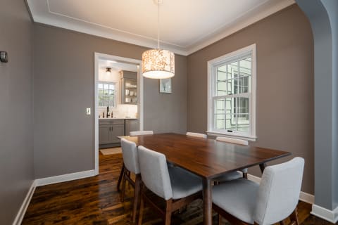 Dining room featuring a wooden table surrounded by light gray chairs, with a pendant light above and a view into a kitchen.