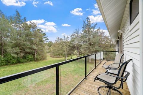A balcony with wooden deck and black chairs, overlooking a grassy field and trees under a blue sky.