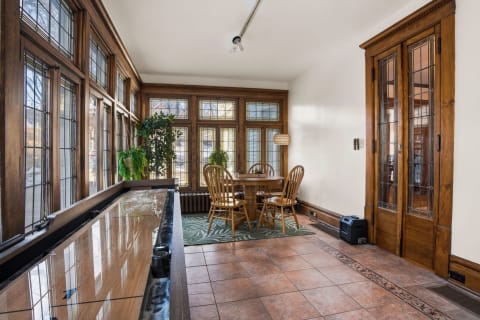A sunlit porch featuring large windows, a round wooden dining table, and decorative plants.
