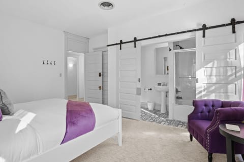A stylish bedroom featuring a white bed with a purple throw, a purple armchair, and a barn-door bathroom.