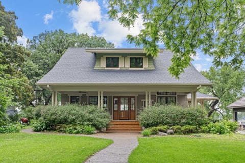 A two-story house with a gray roof and green shutters, surrounded by greenery.