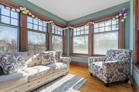 Sunroom featuring a beige sofa, matching armchair, colorful garland, and large windows.