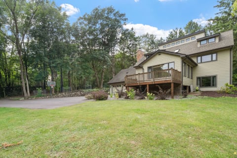 A modern house with a wooden deck, surrounded by trees and a lawn, featuring a basketball hoop in the driveway.
